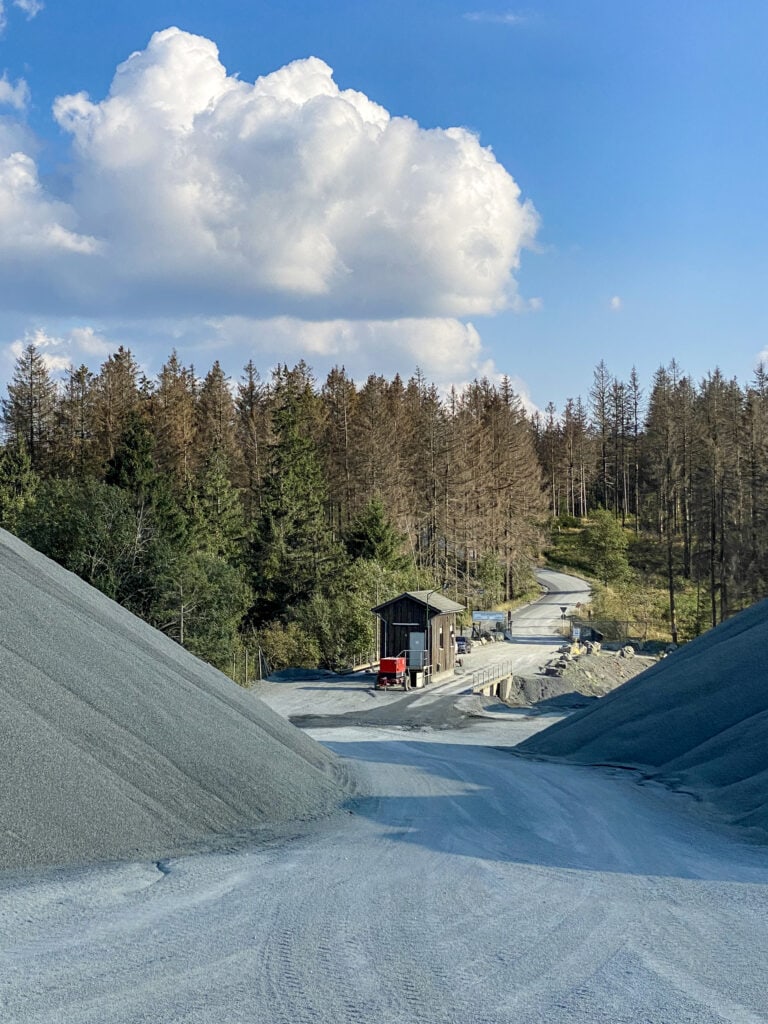 Steinbruch mit großen grauen Kieshügeln, einer Überflurwaage, umgeben von Wald, blauer Himmel mit Wolken, robust und präzise für Wägetechnik geeignet.