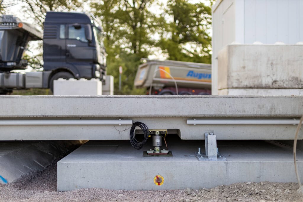 Seitliche Ansicht einer Überflurfahrzeugwaage im Rohbau, eingebettet in Beton. Kabel und Elektronik sichtbar. Im Hintergrund unscharf ein Lkw und grüne Bäume. Robust, präzise, unkompliziert.