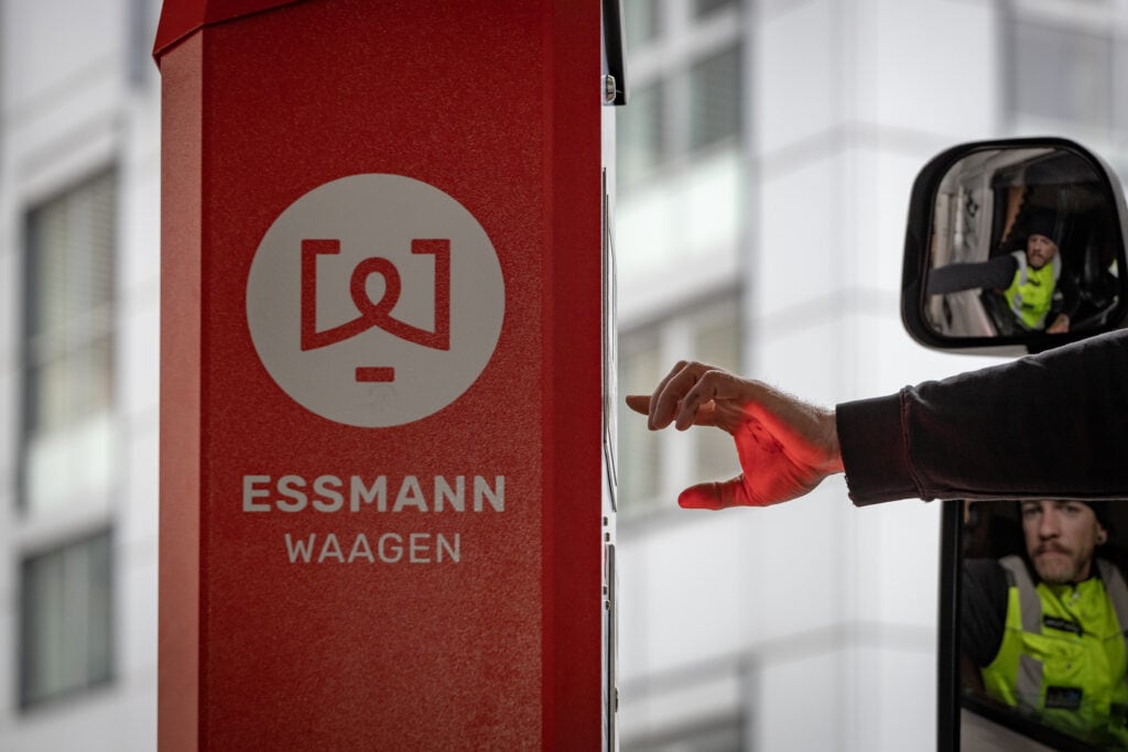 Rote Essmann-Selbstbediensäule mit Logo, Hand bedient sie. Mann in gelber Weste schaut aus Lkw-Spiegel. Modernes, funktionales Umfeld.
