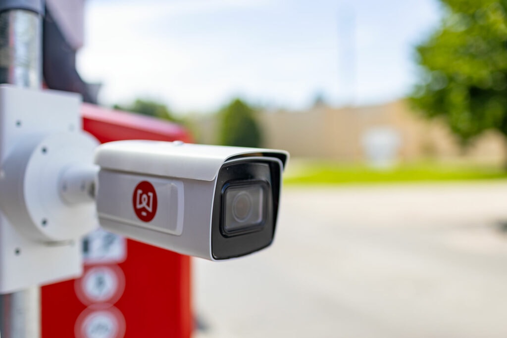 White and silver surveillance camera with logo, mounted on a bracket; blurred background with red object and green tree; license plate recognition theme.