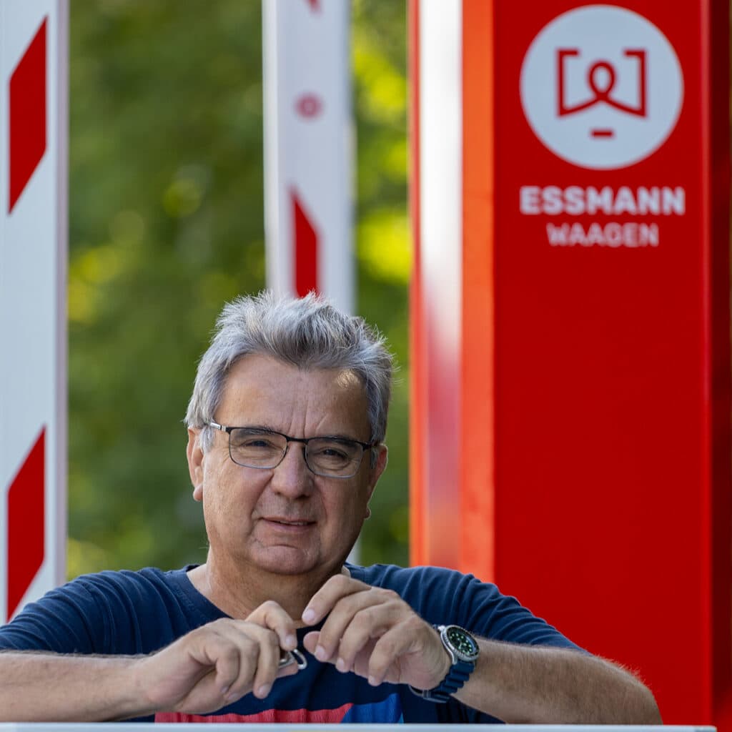 Mann mit grauen Haaren und Brille. Hintergrund zeigt rote Selbstbediensäule mit Aufschrift 'Essmann Waagen'. Umgebung grün, angedeutete Laubbäume. Kontext: Niederlassung Süd.