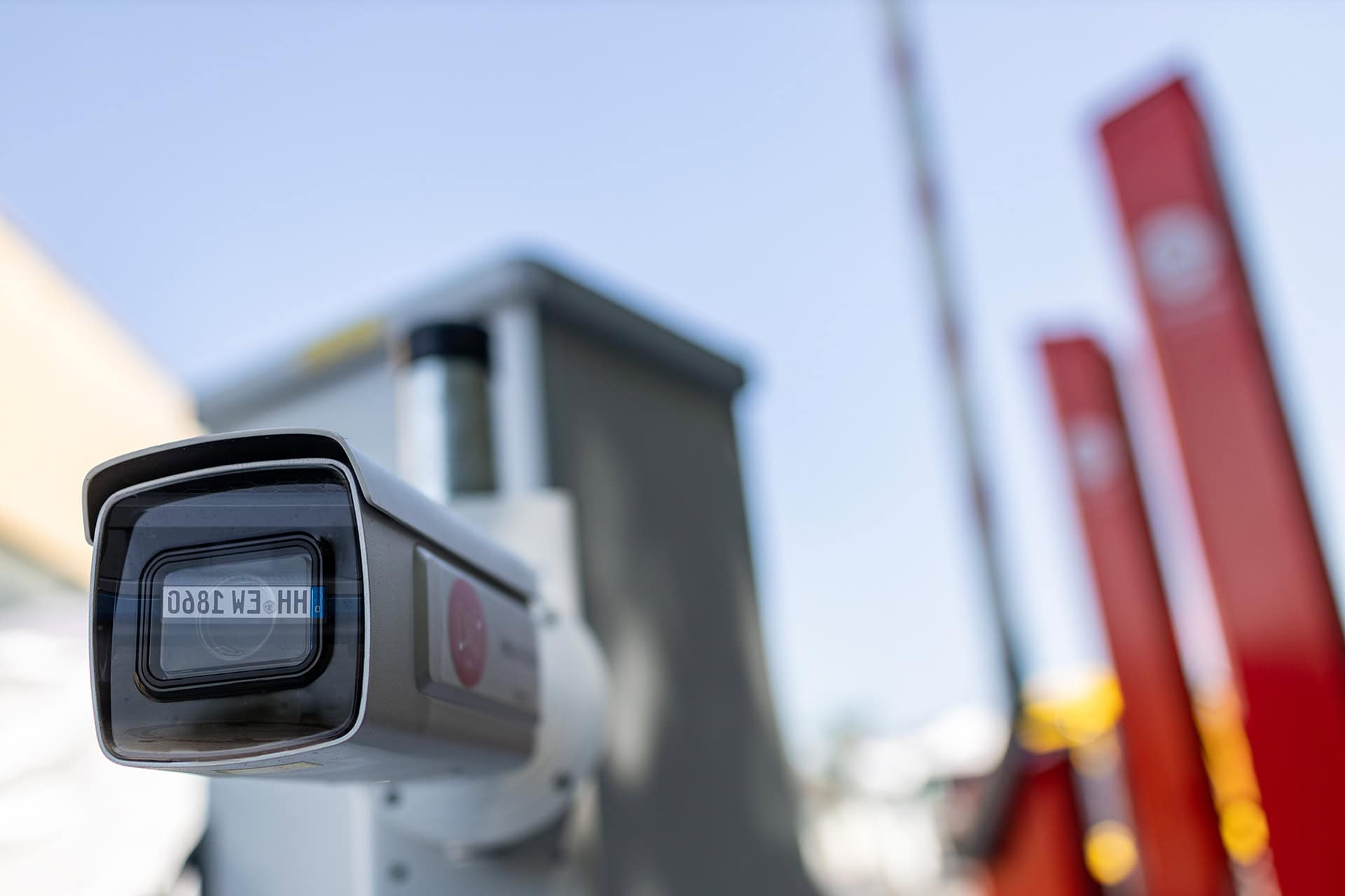 Close-up of a surveillance camera for license plate recognition from ESSMANN. In the background, blurred red barriers and a cloudless sky. Clear, functional impression.