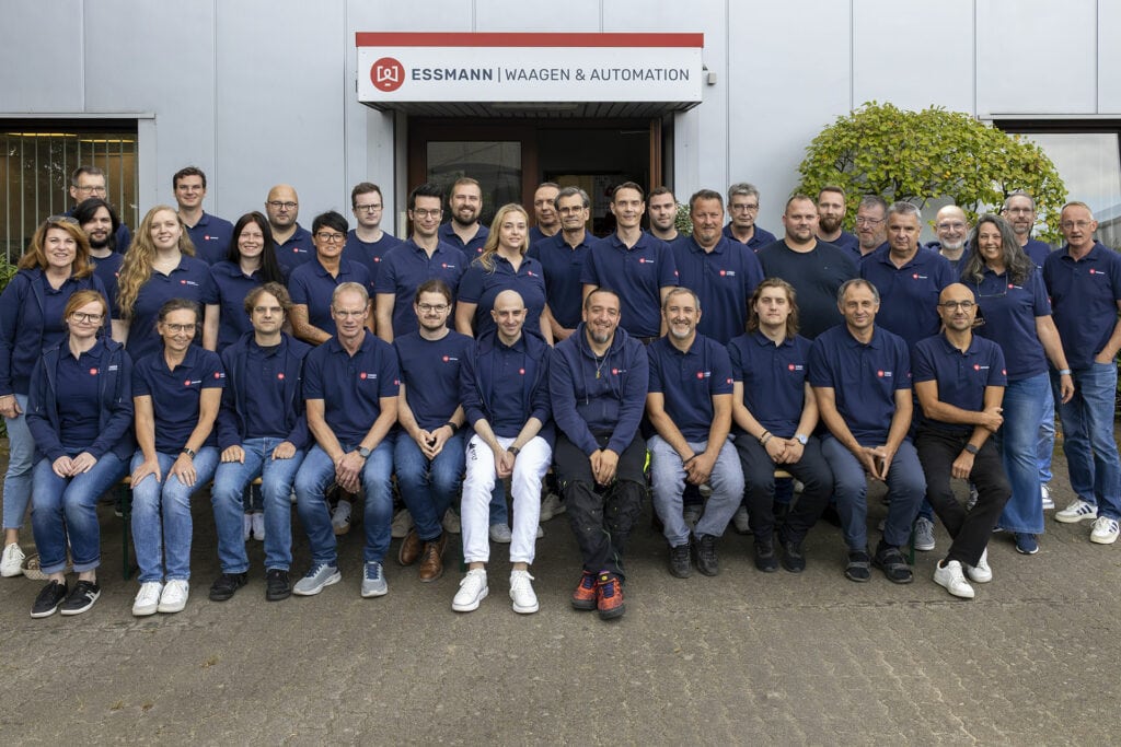 Gruppe von 37 Personen in blauen Polohemden vor einem Gebäude der Essmann Gruppe, lächelnd, einige sitzen auf einer Bank. Gebäude mit Essmann-Logo und Beschriftung 'Waagen & Automation'.