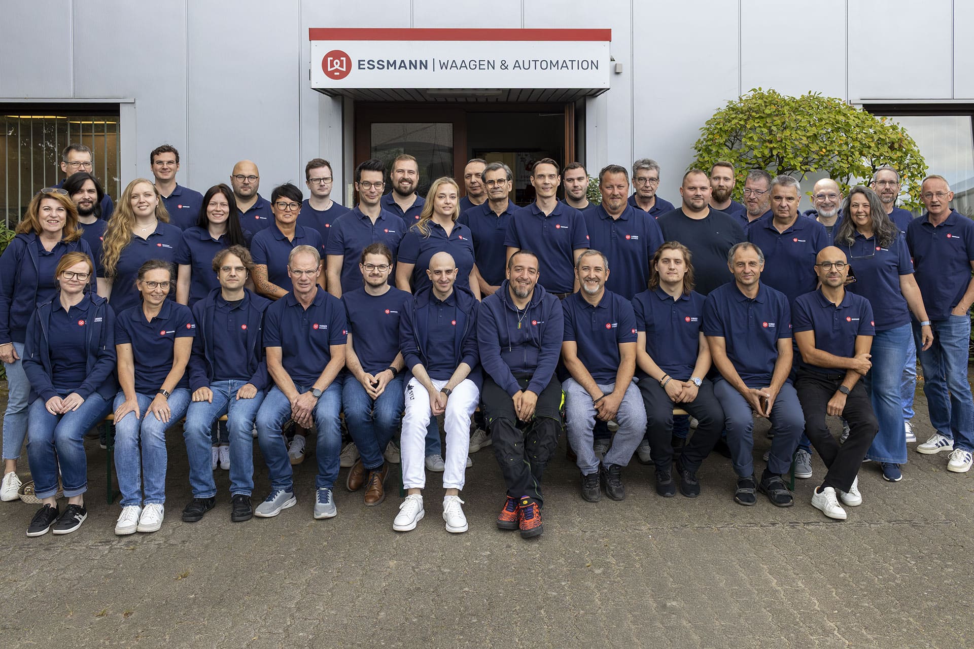 Gruppe von 37 Personen in blauen Polohemden vor einem Gebäude der Essmann Gruppe, lächelnd, einige sitzen auf einer Bank. Gebäude mit Essmann-Logo und Beschriftung 'Waagen & Automation'.