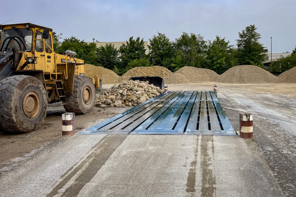 Gelber Radlader neben einer Überflur-Fahrzeugwaage im Rohbau, umgeben von Kies- und Steinbrocken. Im Hintergrund grüne Bäume und Kieshaufen. Robust und zweckmäßig für Steinbruch.
