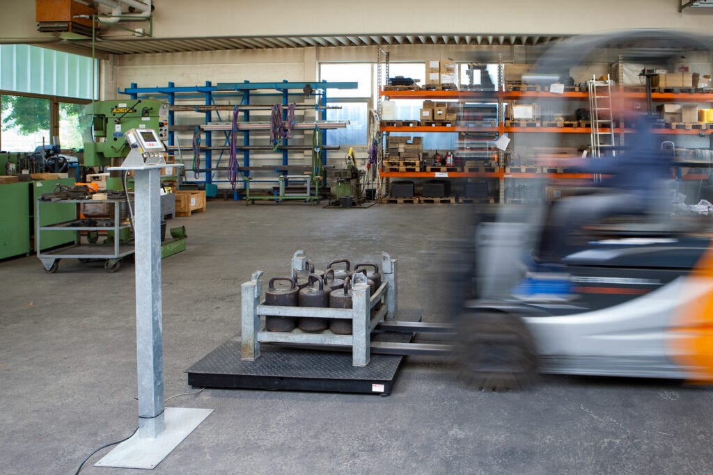 Werkstatt mit Plattformwaage, darauf schwere Gewichte. Gabelstapler in Bewegung. Regale im Hintergrund mit Werkzeugen und Materialien. Industrielle Atmosphäre, Licht fällt durch Fenster.