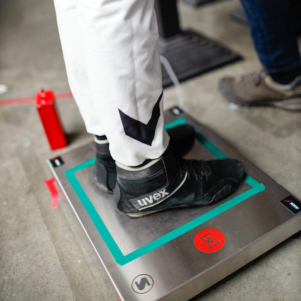 Person in weißer Hose und schwarzen Schuhen steht auf einer silbernen Soehnle Plattformwaag. Grüner Rahmen, rotes Markenlogo, grauer Boden, rote Flasche im Hintergrund.
