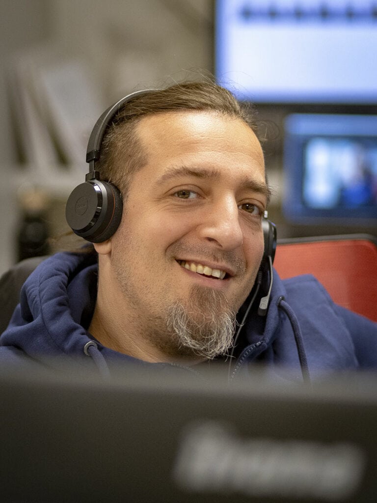 Mann mit Headset und blauem Kapuzenpullover lächelt in die Kamera, im Hintergrund ein unscharfer Bildschirm. Kontext: ESSMANN-IT-Support Dennis im Einsatz für das Technische Hilfswerk.