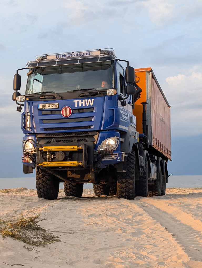 Blauer THW-LKW mit Tatra-Logo und stralsunder Kennzeichen auf sandigem Gelände. Fahrer im Führerhaus. Dahinter orangefarbener Anhänger. Überwiegend sonnig mit teilweise bewölktem Himmel.
