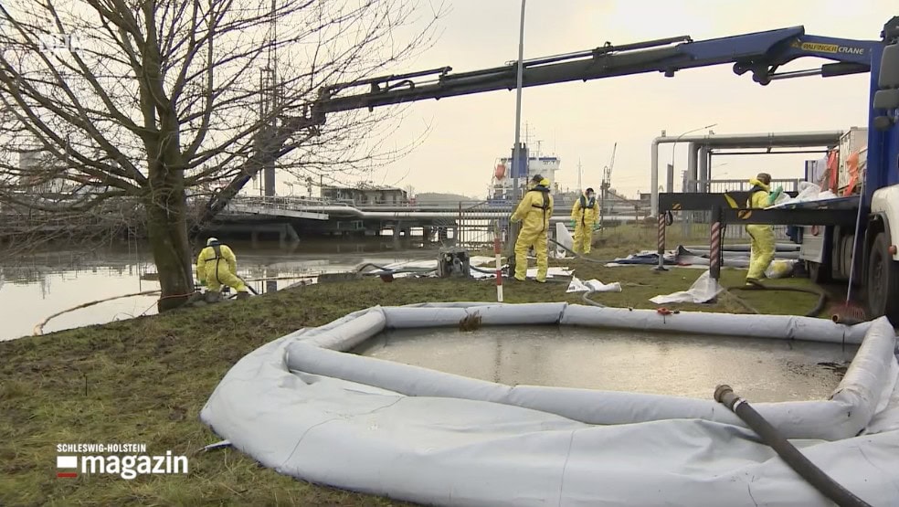Drei Personen in gelben Schutzanzügen arbeiten am Ufer eines Gewässers. Ein Lastwagen mit Kran steht daneben. Im Vordergrund ein aufblasbares Becken. Industrieanlagen im Hintergrund.