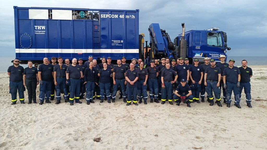 Gruppe von THW-Helfern in blau-schwarzer Uniform steht vor großem blauen Fahrzeug mit Aufschrift 'THW Fachgruppe Ölschaden', am Strand mit Meer im Hintergrund.