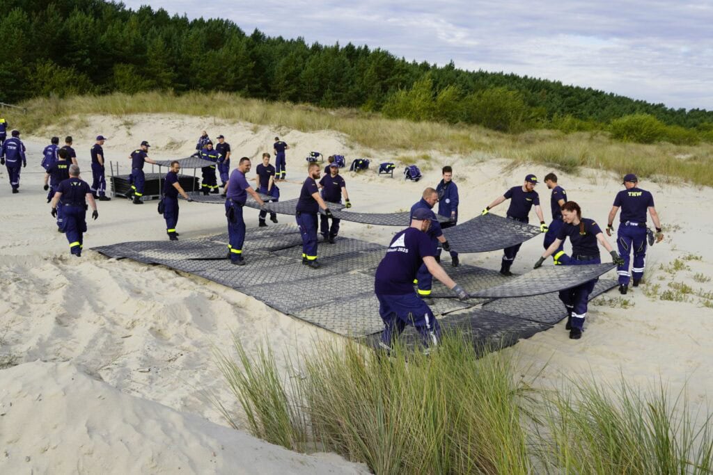 Gruppe von Menschen in blauen THW-Uniformen legt Matten auf sandigem Boden. Sie arbeiten gemeinsam auf einer offenen Fläche mit grasbedeckten Dünen im Hintergrund.