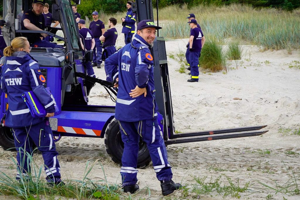 Mehrere Personen in THW-Uniformen arbeiten zusammen auf einem sandigen Gelände mit einem Gabelstapler. Gräser ringsherum, entspannte Stimmung. Foto: THW/Claus Böttcher