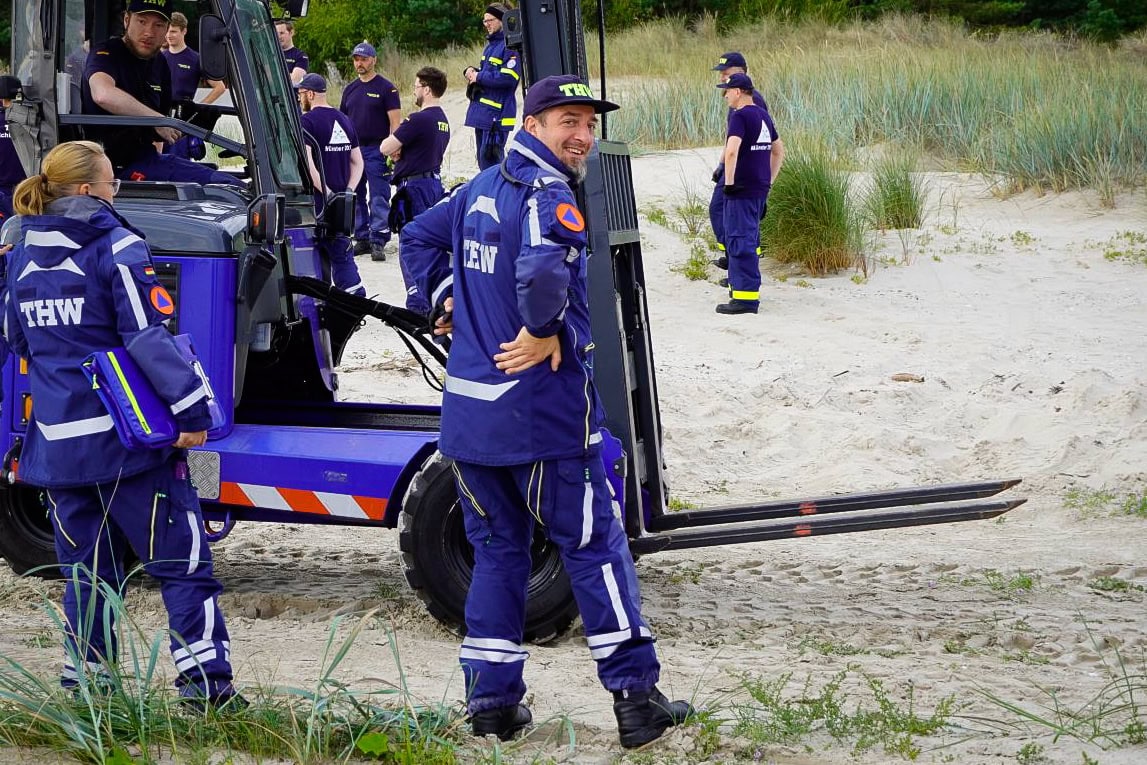 Mehrere Personen in THW-Uniformen arbeiten zusammen auf einem sandigen Gelände mit einem Gabelstapler. Gräser ringsherum, entspannte Stimmung. Foto: THW/Claus Böttcher