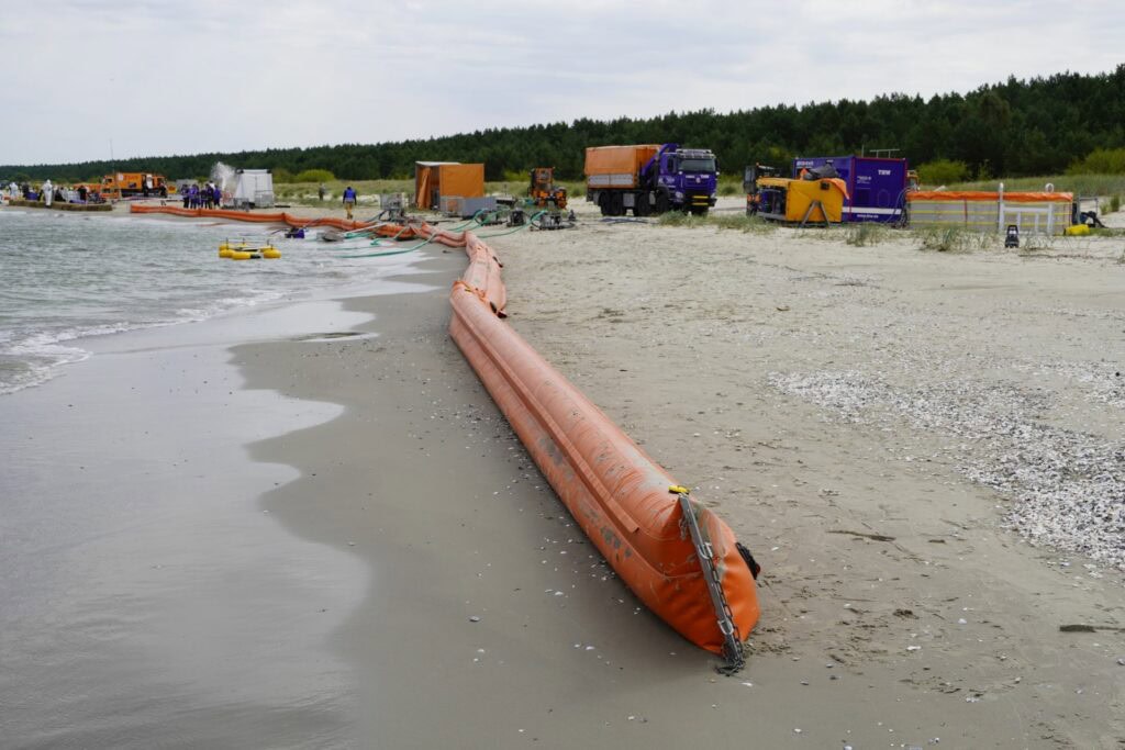 Am Strand liegt eine lange orangefarbene Ölsperre zum Meer. Im Hintergrund sind THW-Fahrzeuge, Zelte und Personen. Der Himmel ist grau, der Strand sandig und teils bewachsen.