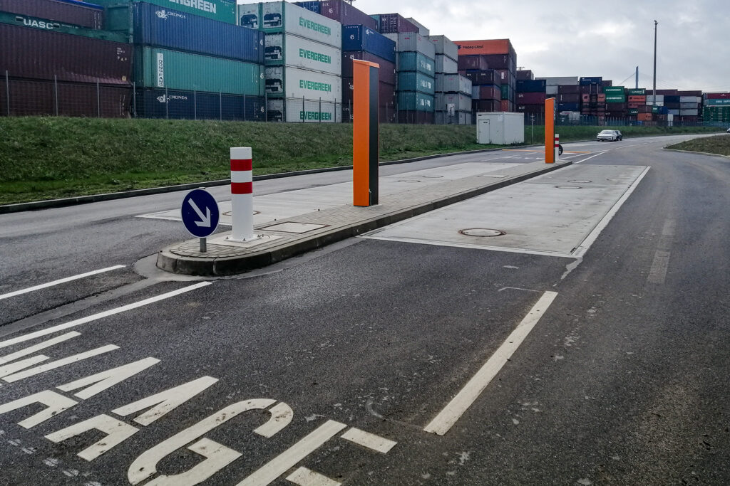 Unterflur-Fahrzeugwaage auf Straße, weiße Markierungen, blaue Richtungsschilder, orange Posten. Containerstapel im Hintergrund, grauer Himmel, Auto in der Distanz, Containerterminal-Atmosphäre.