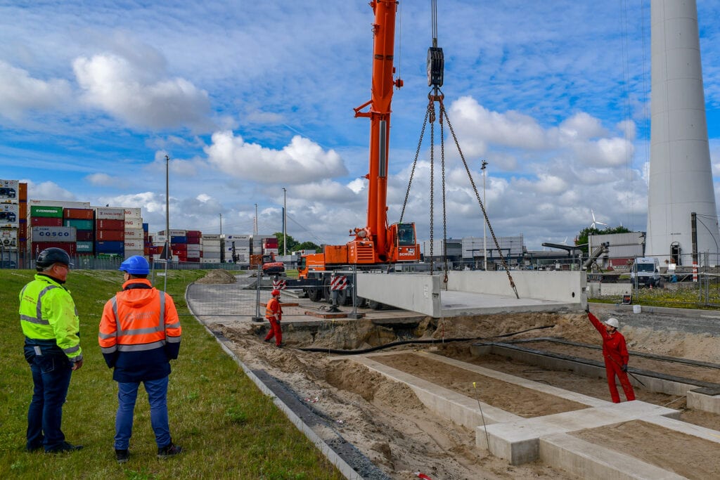 Zwei Männer in Warnkleidung blicken auf Arbeiter und orange Kran, der Betonplatte auf Baustelle platziert. Containerstapel, blauer Himmel mit Wolken im Hintergrund. Titel: 2022 Hamburg.