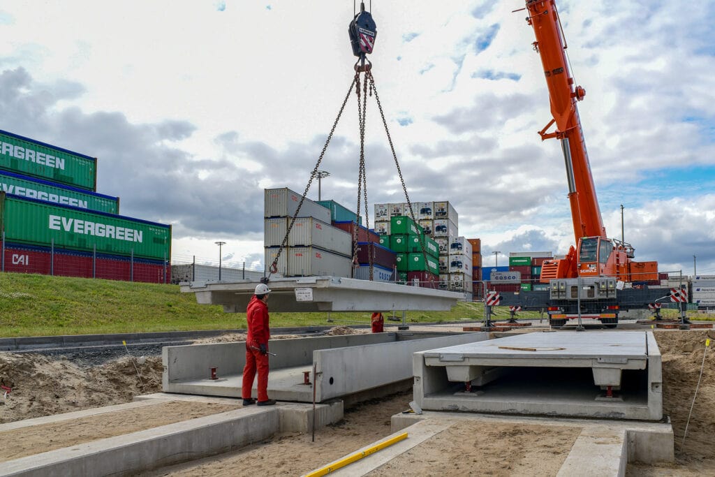 Kran hebt große Betonplatte über Baugrube, Arbeiter in roter Kleidung überwacht. Hintergrund: gestapelte Container. Thema: Präzision bei schwimmendem Baugrund in Hamburg-Hafen.
