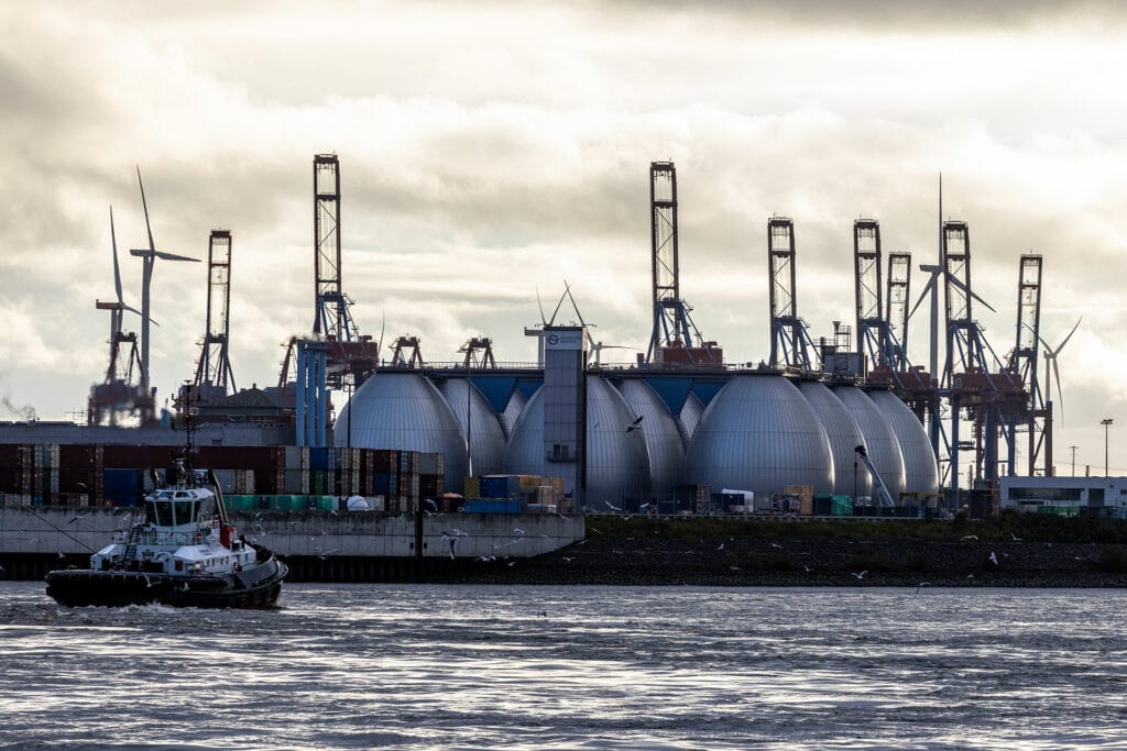 Industriegelände am Wasser mit mehreren großen, kuppelförmigen Anlagen. Im Hintergrund befinden sich Kräne und Windräder. Ein Schlepperschiff fährt auf dem Wasser, Möwen fliegen umher.