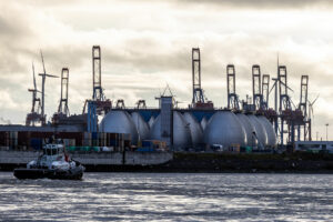 Industriegelände am Wasser mit mehreren großen, kuppelförmigen Anlagen. Im Hintergrund befinden sich Kräne und Windräder. Ein Schlepperschiff fährt auf dem Wasser, Möwen fliegen umher.