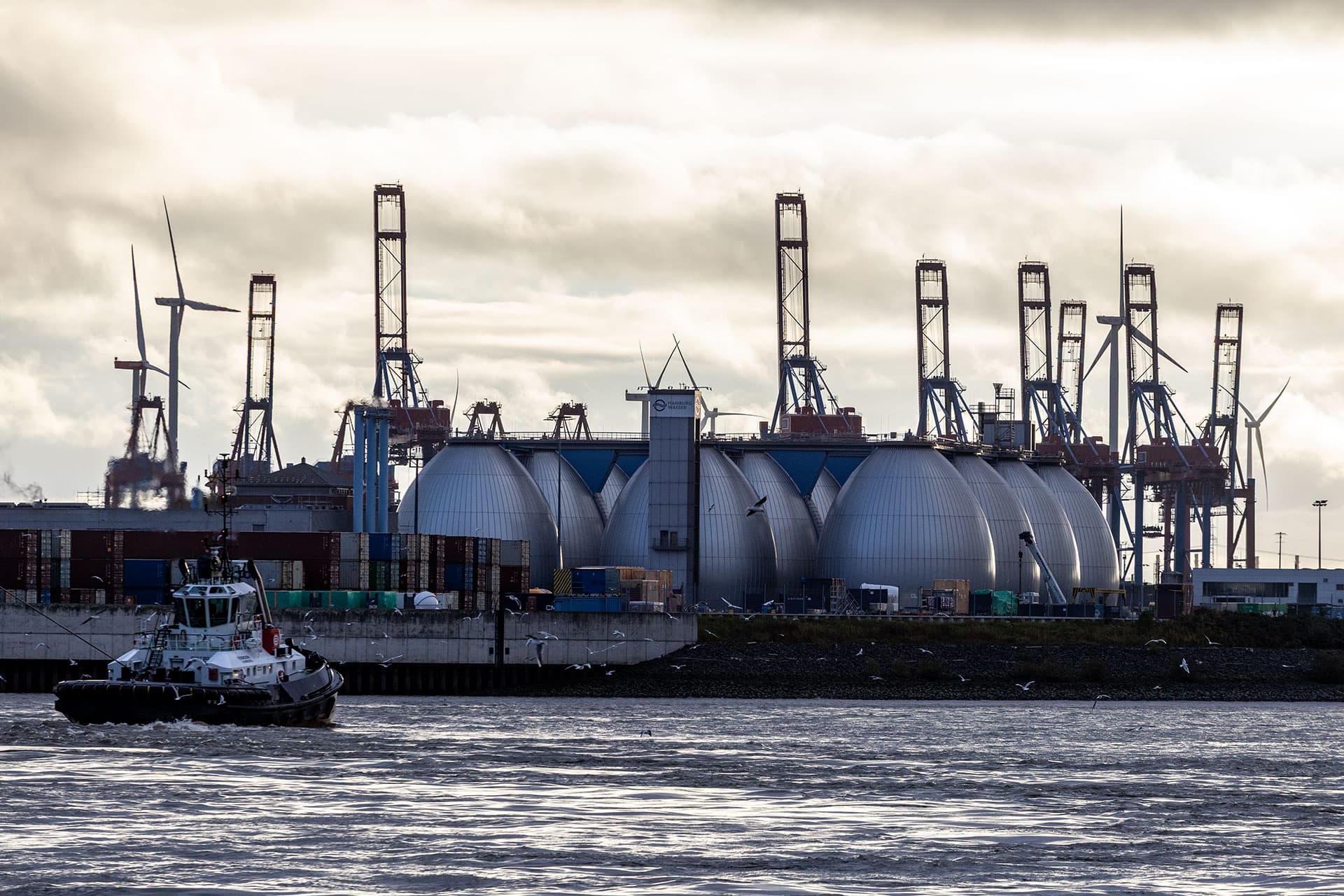 Industriegelände am Wasser mit mehreren großen, kuppelförmigen Anlagen. Im Hintergrund befinden sich Kräne und Windräder. Ein Schlepperschiff fährt auf dem Wasser, Möwen fliegen umher.
