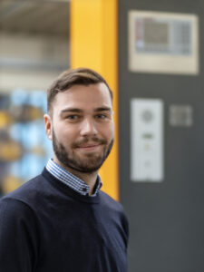 Ein junger Mann mit Bart lächelt, trägt dunkelblauen Pullover, kariertes Hemd. Hinter ihm unscharf eine orange-graue Selbstbediensäule.