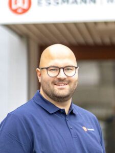 Lächelnder Mann mit Glatze und Brille in einem ESSMANN-Shirt vor dem Hauptgebäude