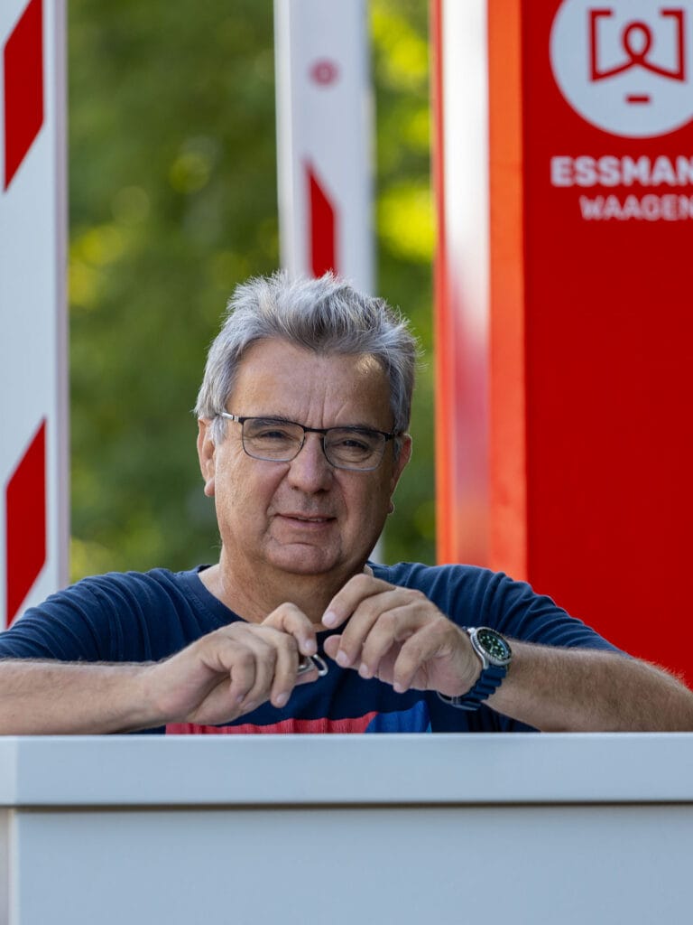 Ein weiser Mann mit grauen Haaren und Brille steht draußen vor einer rot-weißen Schranke. Im Hintergrund ist eine rote Selbstbediensäule mit der Aufschrift 'ESSMANN WAAGEN'. Er schaut ruhig in die Kamera.