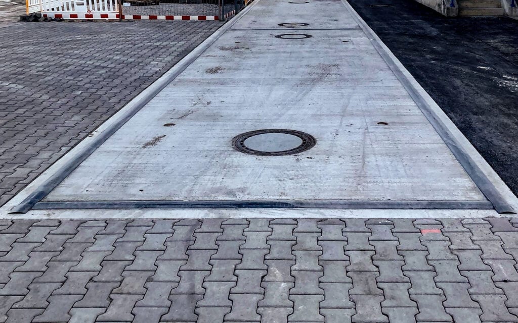 Fahrzeugwaage aus Beton mit runden Metallabdeckungen in einer gepflasterten Fläche. Im Kontext Rhenus Logistics, robuste Verwiegung mit HE-SFK 20 WHG, Umgebung in grau und schwarz.