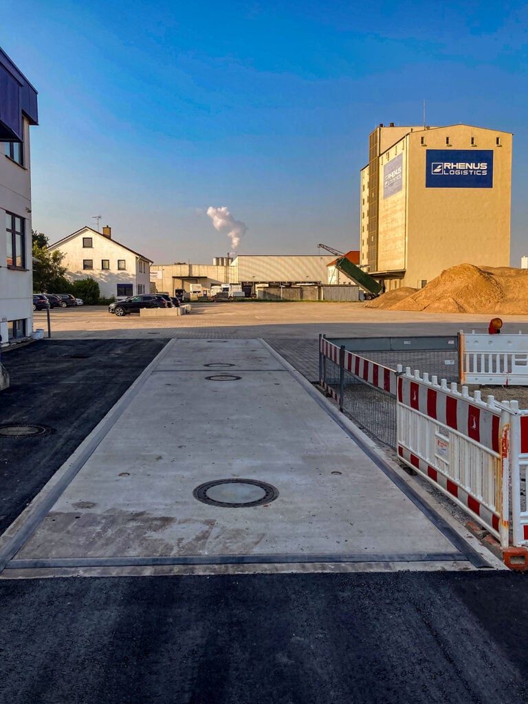 Industriegelände bei Rhenus Logistics mit Fahrzeugwaage HE-SFK 20 WHG im Vordergrund. Im Hintergrund sind Lagergebäude, Fabrikschornstein und ein blauer Himmel sichtbar.