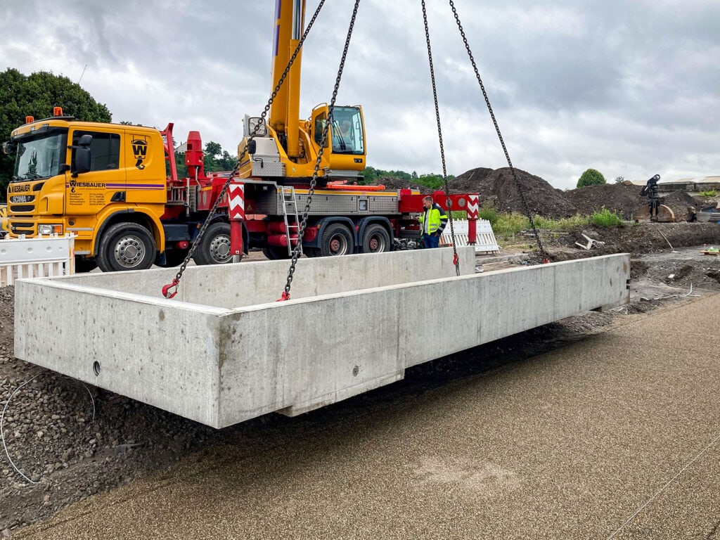 Gelber Kranwagen hebt Betonfundament an Ketten. Zwei Männer tragen Warnwesten, gelber LKW im Hintergrund. Baustelle mit Erde und Vegetation, bewölkter Himmel.