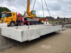 Gelber Kranwagen hebt Betonfundament an Ketten. Zwei Männer tragen Warnwesten, gelber LKW im Hintergrund. Baustelle mit Erde und Vegetation, bewölkter Himmel.