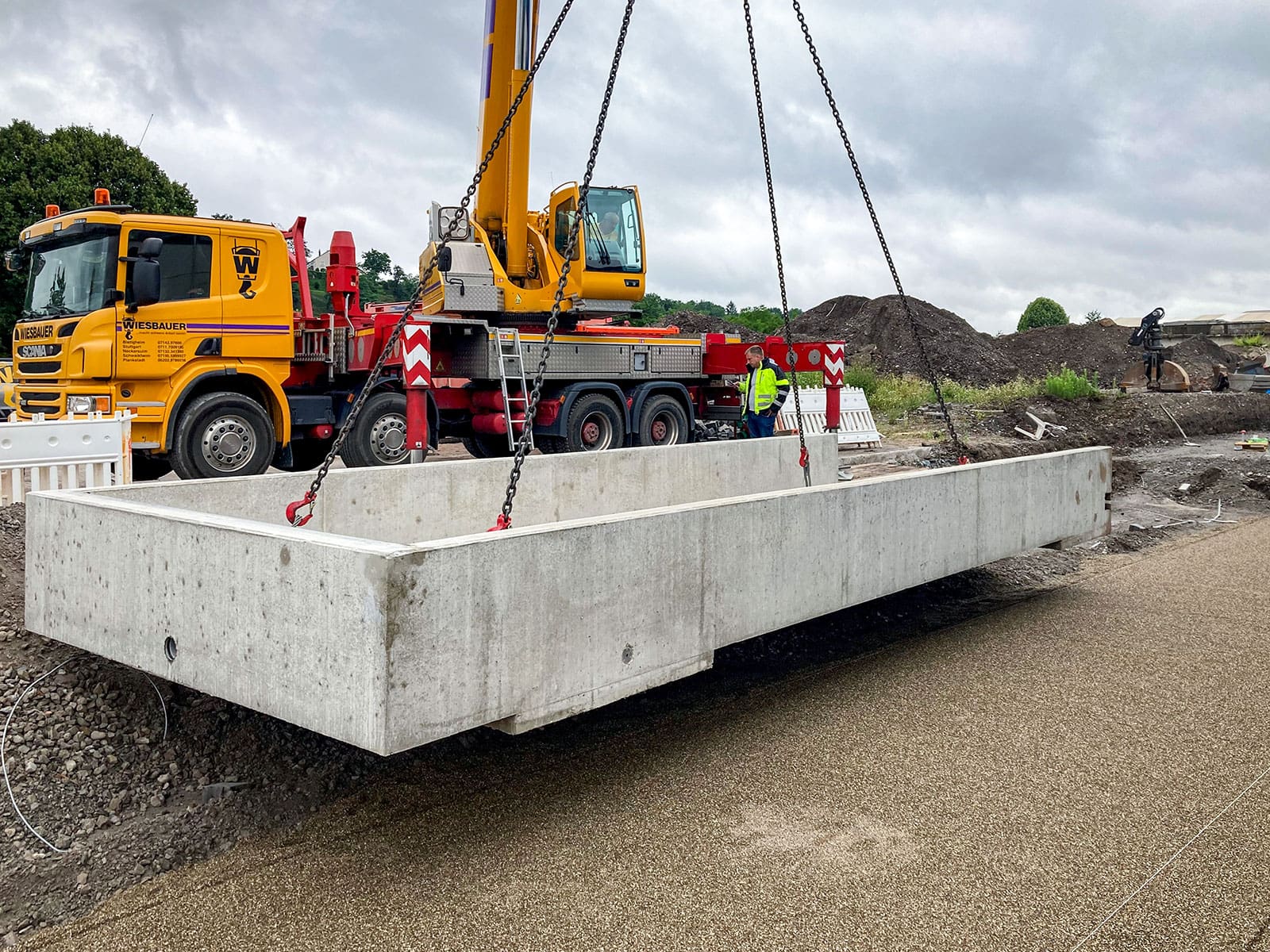 Gelber Kranwagen hebt Betonfundament an Ketten. Zwei Männer tragen Warnwesten, gelber LKW im Hintergrund. Baustelle mit Erde und Vegetation, bewölkter Himmel.