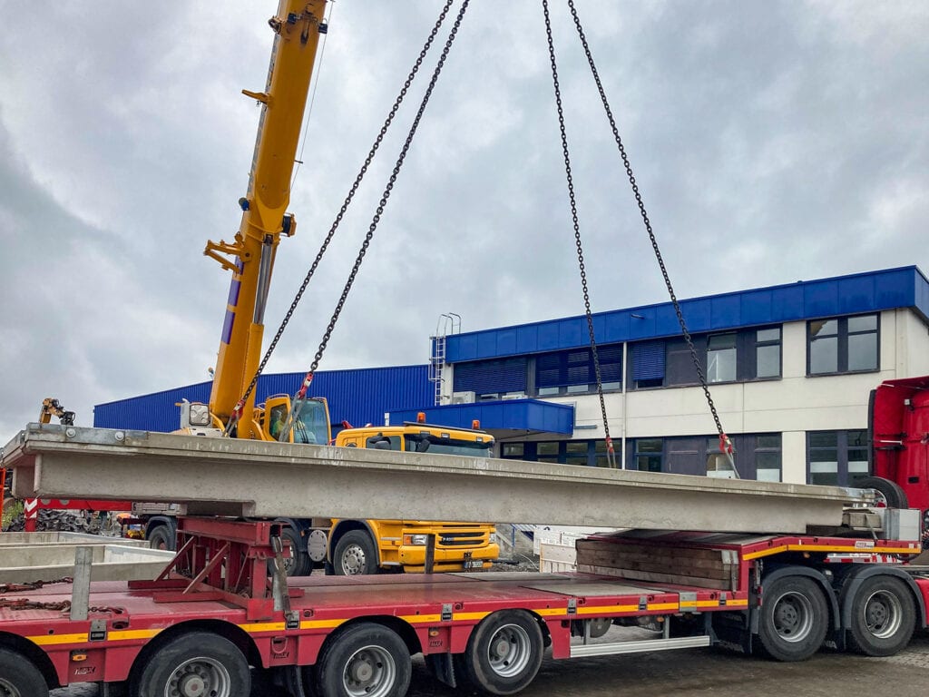 Gelber Kran hebt Betonträger auf roten Tieflader. Im Hintergrund blau-weißes Gebäude unter grauem Himmel. Einsatz bei Rhenus Logistics für Verwiegung mit HE-SFK 20 WHG Fahrzeugwaage.