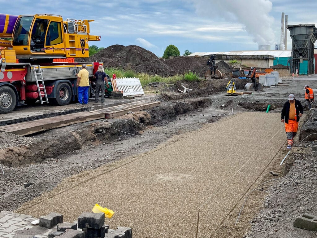 Baustelle mit Bauarbeiter in orangefarbenen Warnwesten, die eine gerade Fläche aus Kies ebnen. Gelber Lastwagen mit Kran auf der linken Seite, Bagger im Hintergrund. Wolken am Himmel.
