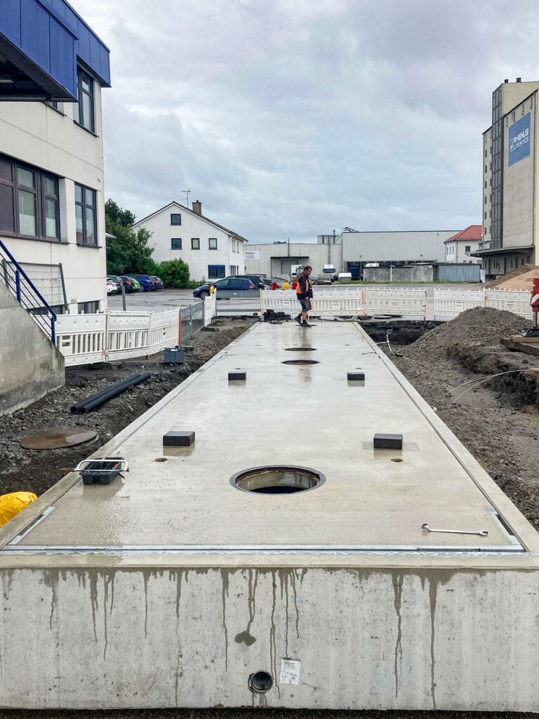 Baustelle mit großer Betonplatte, möglicherweise eine Fahrzeugwaage, umgeben von Absperrungen. Eine Person mit Warnweste steht darauf. Im Hintergrund sind Gebäude und parkende Autos sichtbar.