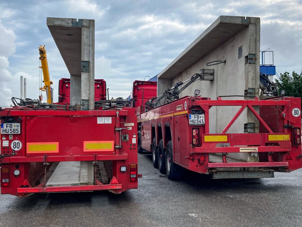 Zwei rote Schwertransporter mit Betonplatten, gelber Kran im Hintergrund, bewölkter Himmel. Rhenus 7: Robuste Verwiegung bei Rhenus Logistics mit Fahrzeugwaage HE-SFK 20 WHG.