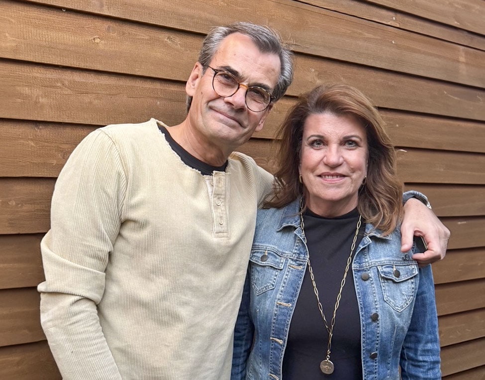 Holger Thiel und Barbara Visser-Thiel lächeln vor einer braunen Holzwand. Links eine Person in beigem Oberteil, rechts eine Person in Jeansjacke und schwarzem Oberteil mit Halskette. Sie wirken freundlich und entspannt.