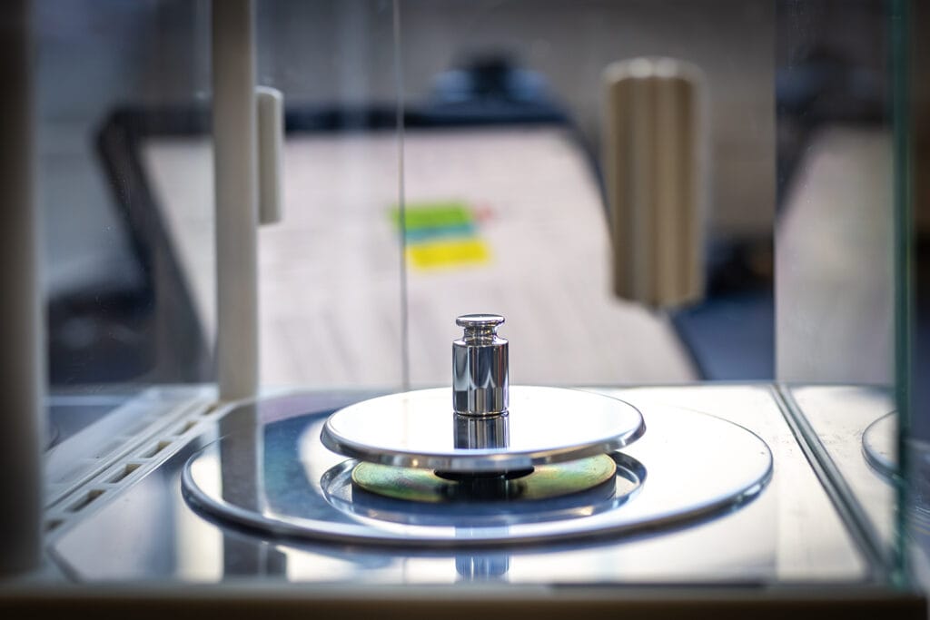 essmann waagenwerkstatt Silbernes Gewicht auf einer Präzisionswage mit gläsernem Windschutz. Weiche Hintergrundunschärfe, im Hintergund ein Tablet mit Kalibrierprotokoll, reflektierte Farben auf der Waage. Essmann Waagenwerkstatt-Umgebung.