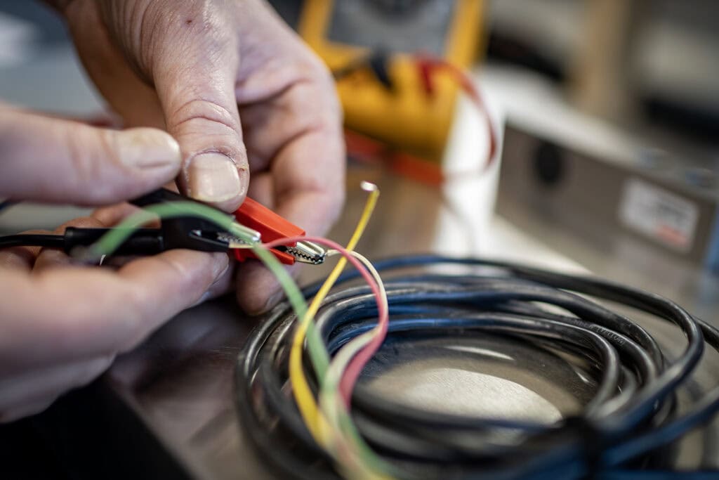 ESSMANN scales workshop Two hands hold a red clip that fixes green, yellow and red wires. Black cable is wound up. A load cell is in the background. Background blurred, yellow and gray recognizable. Context: ESSMANN scale maintenance.