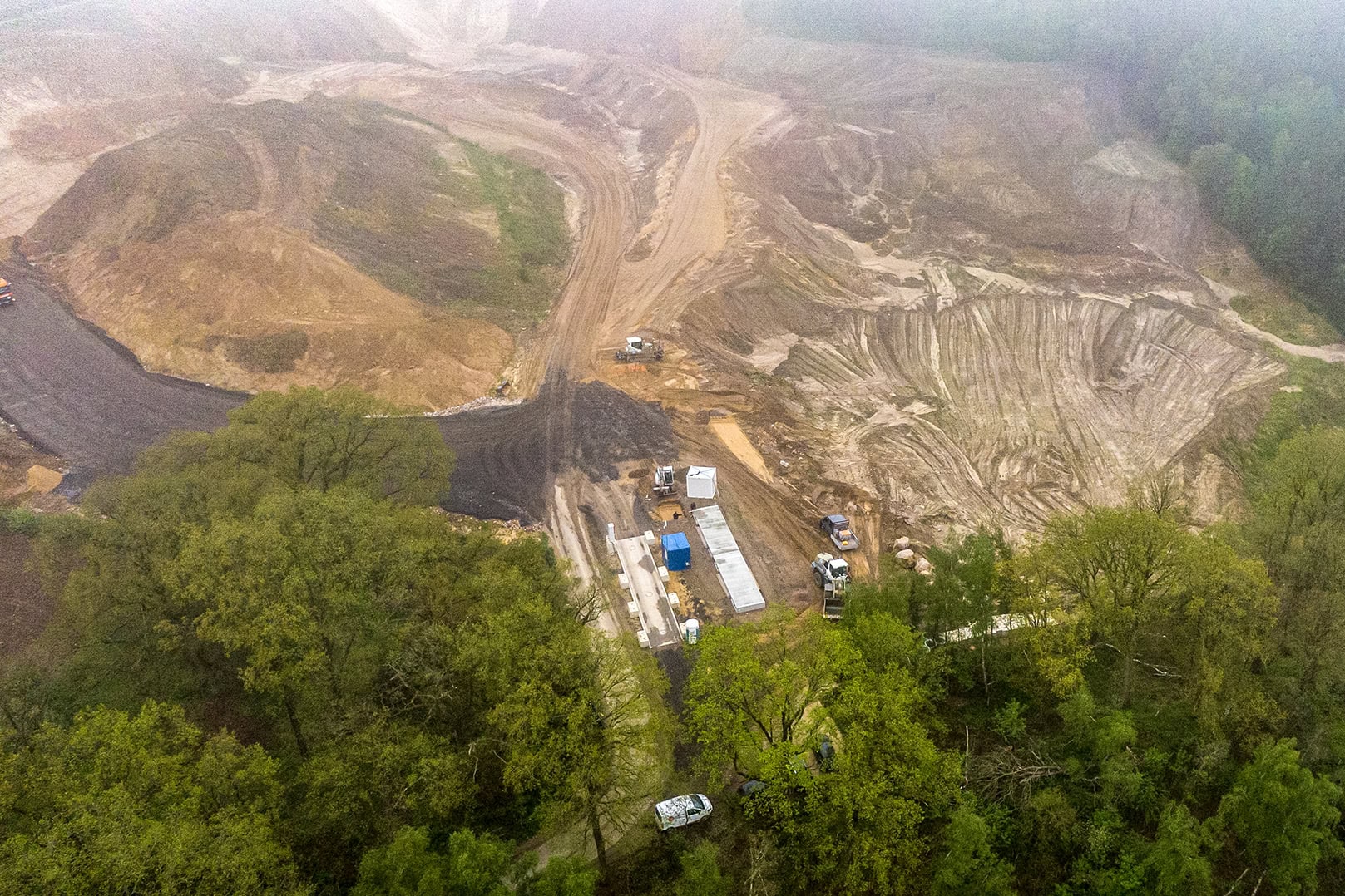 Luftbild eines Steinbruchs und Kieswerks. Erdige Flächen, Gruben und Sandwege. Im Vordergrund grüne Bäume. Mehrere Fahrzeuge und Container auf sandigem Boden. Zengtral sind zwei Fahrzeugwaagen zu sehen.